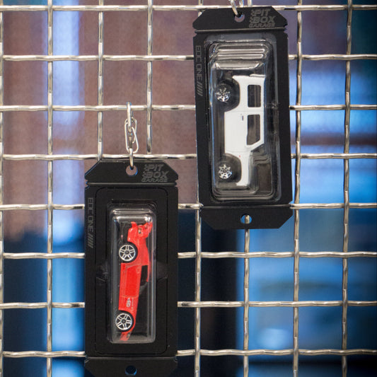 Two miniature die-cast toy cars in black hanging display tags, one red and one white, photographed against a wire grid with a blue reflective background.