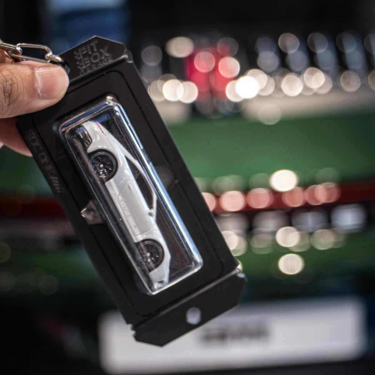 A hand holds a black keychain featuring a miniature silver sports car in a clear plastic window, with a blurred stadium or event background.
