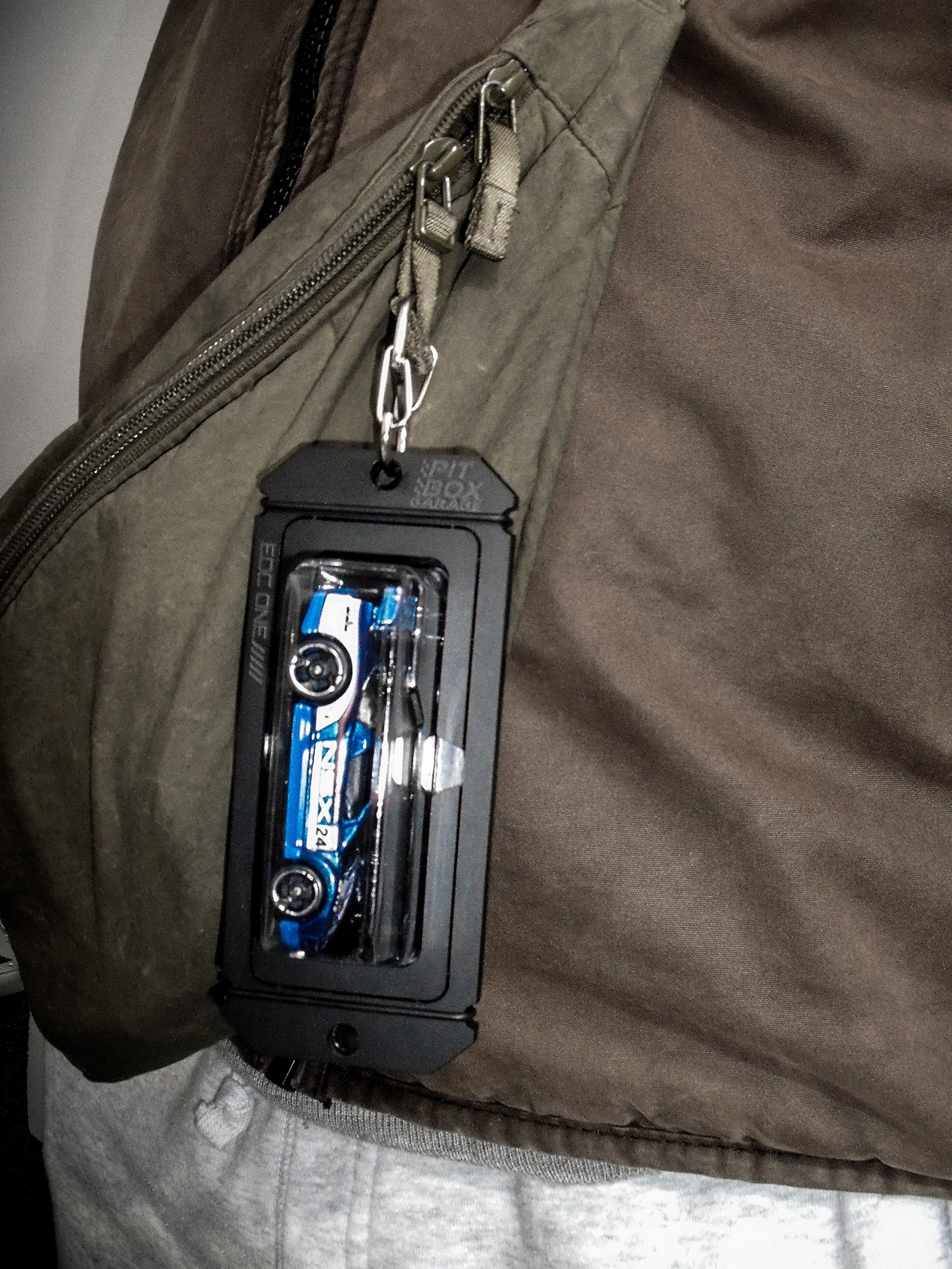 A close-up of a person wearing a brown crossbody bag with a small black rectangular accessory clipped to the zipper. The accessory appears to contain a blue toy car and small white tools inside.