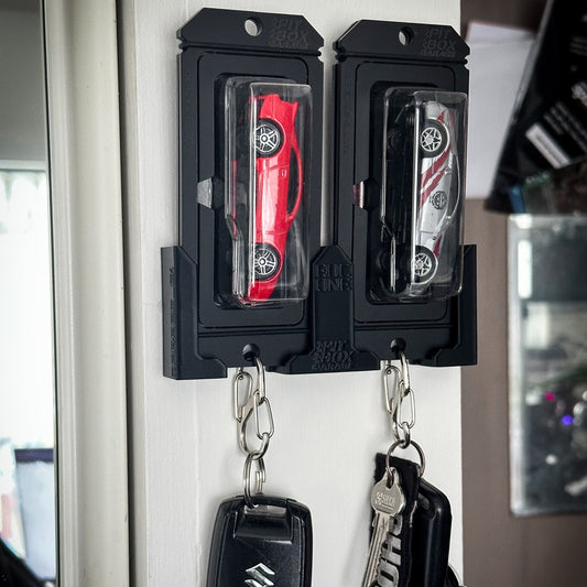 Two black plastic car key holder wall mounts attached to a white door, each holding a car key fob and keys. The left holder contains a red car key, and the right holder contains a silver and white car key.
