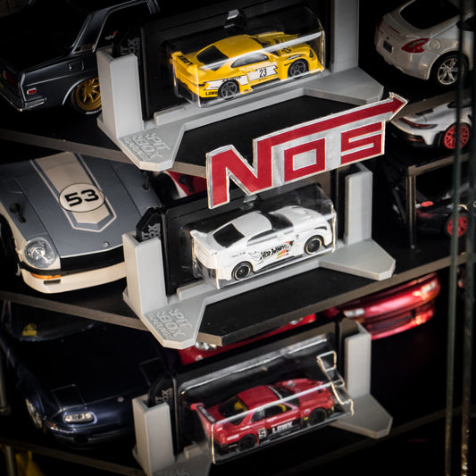 A close-up of a multi-tiered NOS display shelf holding several die-cast model cars, including a yellow race car, a white sports car, a red car, and other cars partially visible on the shelves.