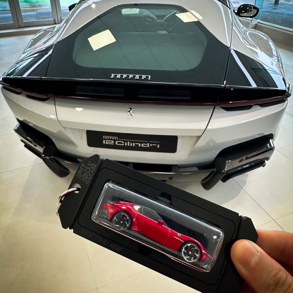 A hand holds a black key fob with a clear window displaying a small red toy sports car, positioned in front of a real-world showroom background featuring a white Ferrari 12 Cilindri rear.