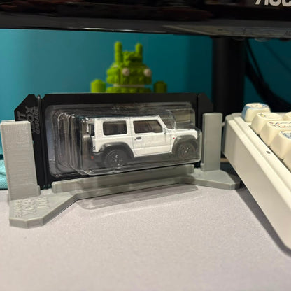 A white die-cast toy SUV in clear blister packaging, displayed on a gray stand on a desk next to a cream-colored mechanical keyboard.