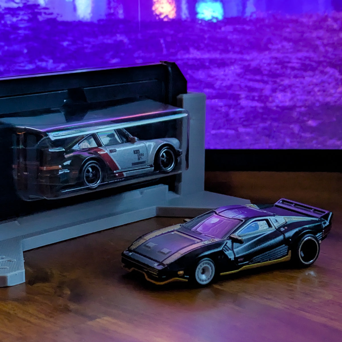 A black die-cast sports car model displayed on a wooden surface in front of a gray display stand holding a silver and red model car, with a purple-lit blurred background.