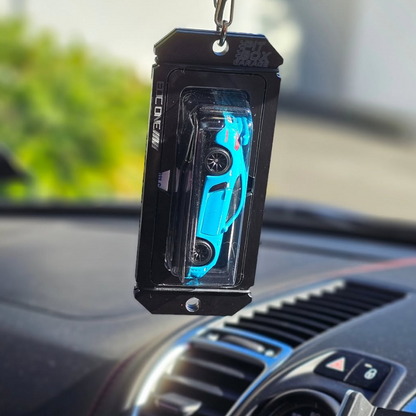 A black car air freshener or keychain holder containing a small blue toy car, hanging from a car's rearview mirror above the dashboard.