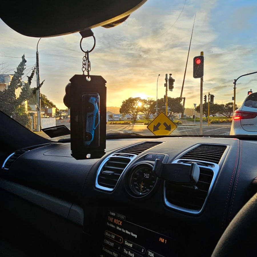 A black car air freshener holder with a blue and black dog figure, hanging from the rearview mirror in a car dashboard at sunset.